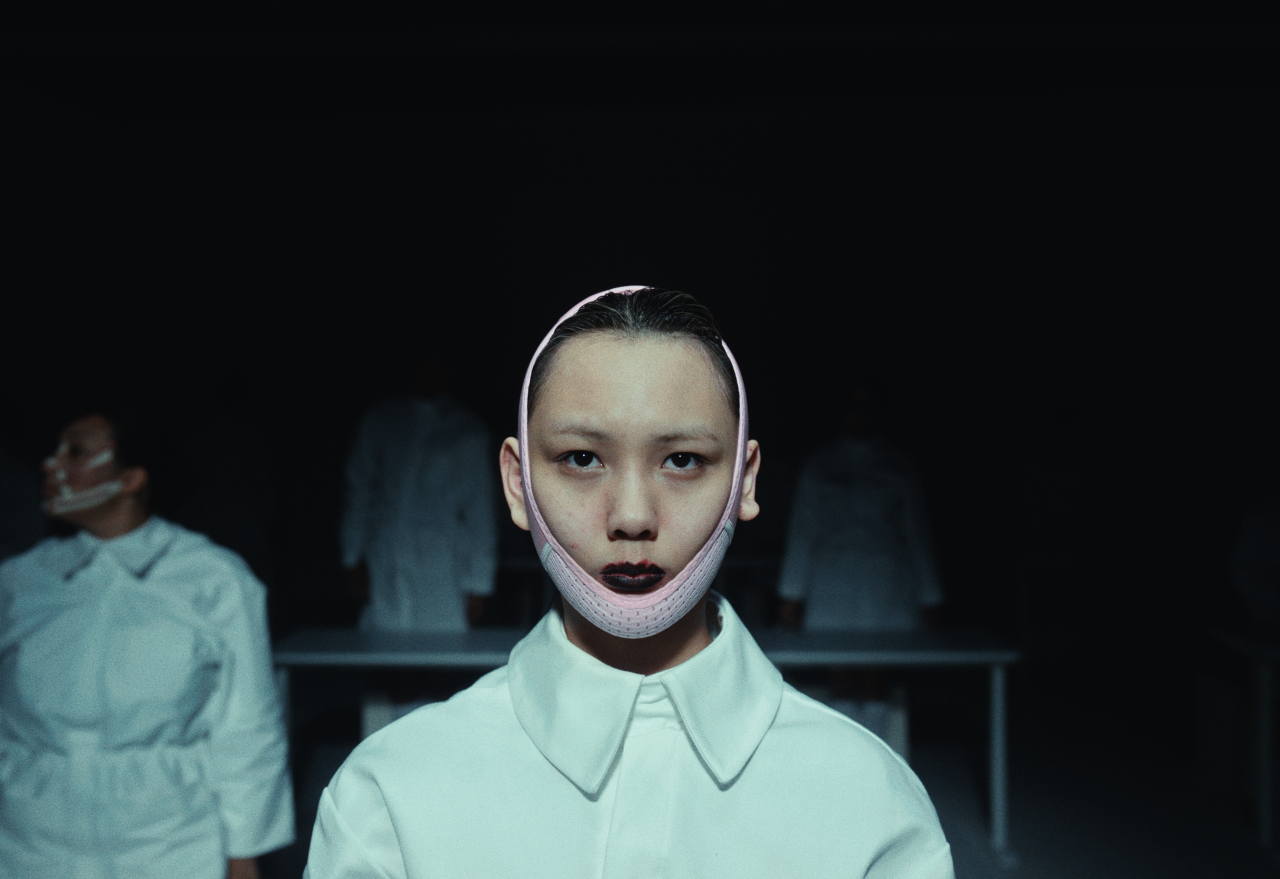 Model wearing white collared shirt with pale makeup and bandaged jaw against dark background in beauty industry parody