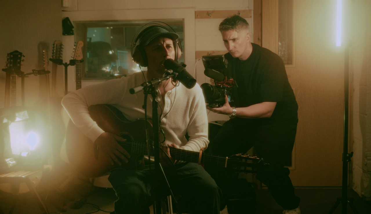 Two people in a studio setting with professional camera and lighting equipment, one wearing headphones and seated while the other stands beside them during filming