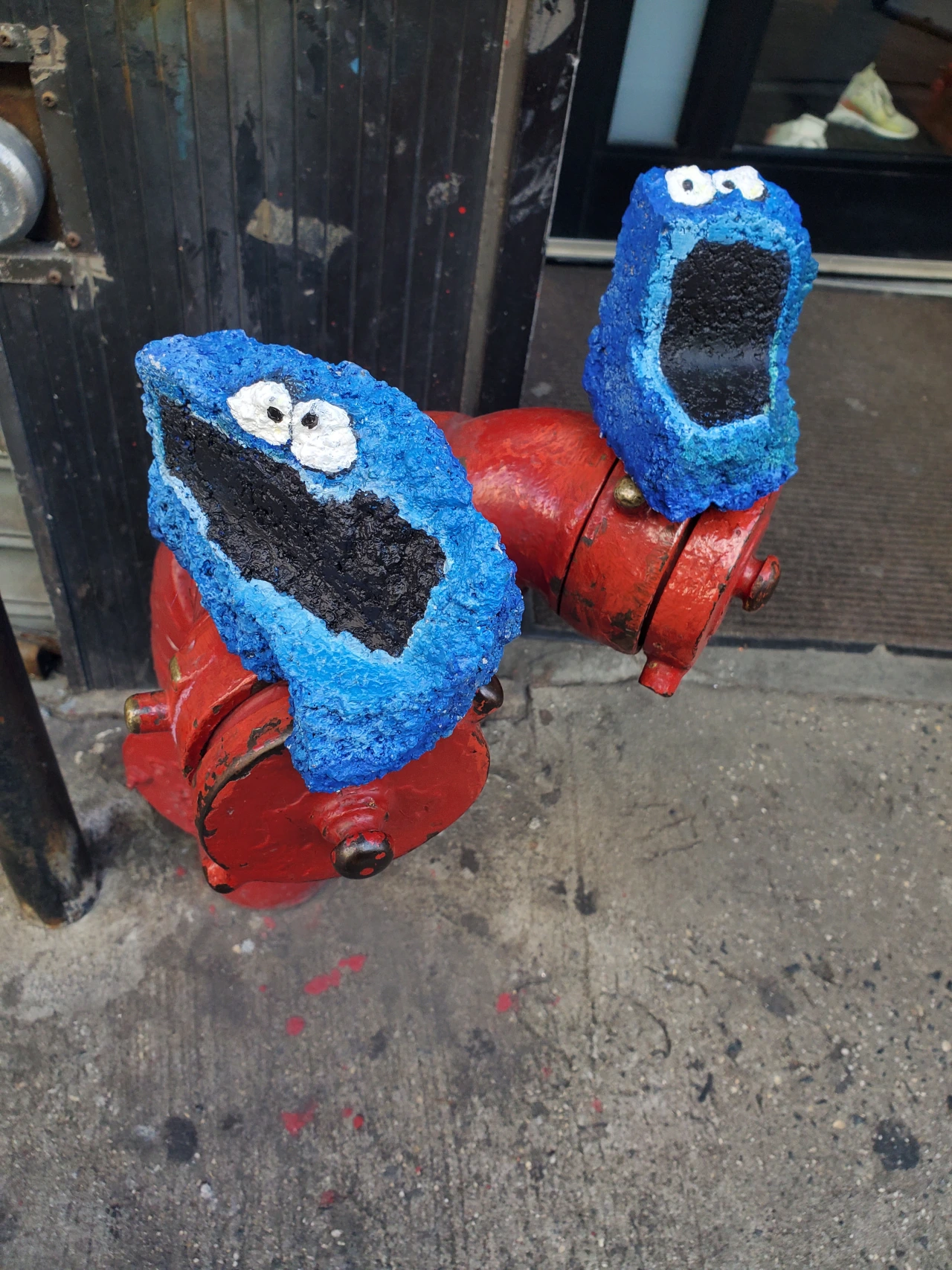 Two painted rocks with blue textured bodies, white eyes and black mouths resembling Cookie Monster characters, mounted on red metal stands on a pavement