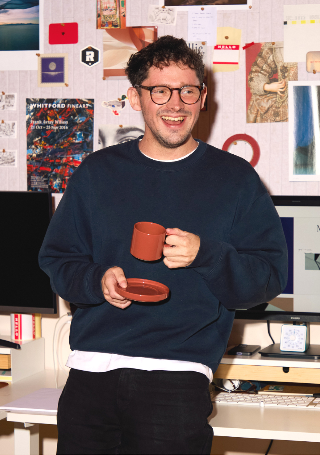 Man wearing glasses and dark sweatshirt holding a red mug and saucer, smiling in front of a mood board covered with sketches, prints and design references