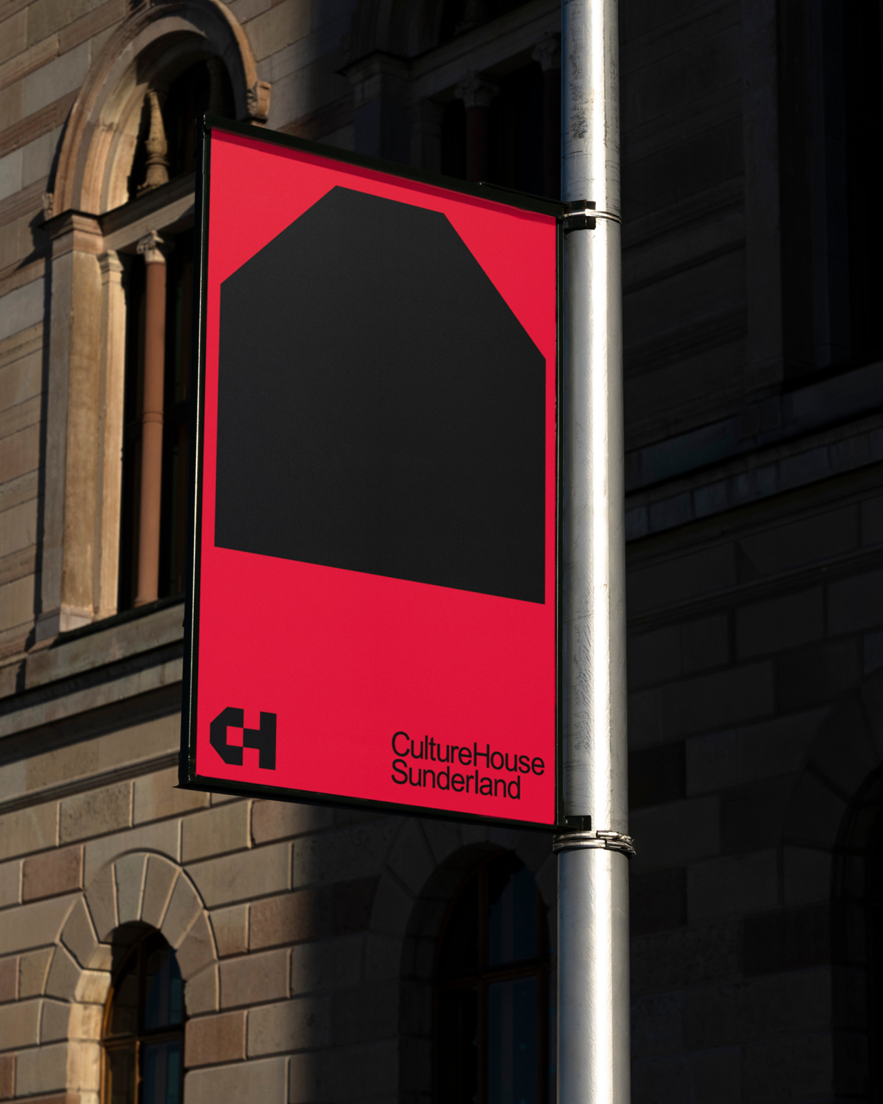 Red and black banner for Culture House Sunderland mounted on a metal pole against a stone building, featuring the CH logo and a house symbol made from folded letters