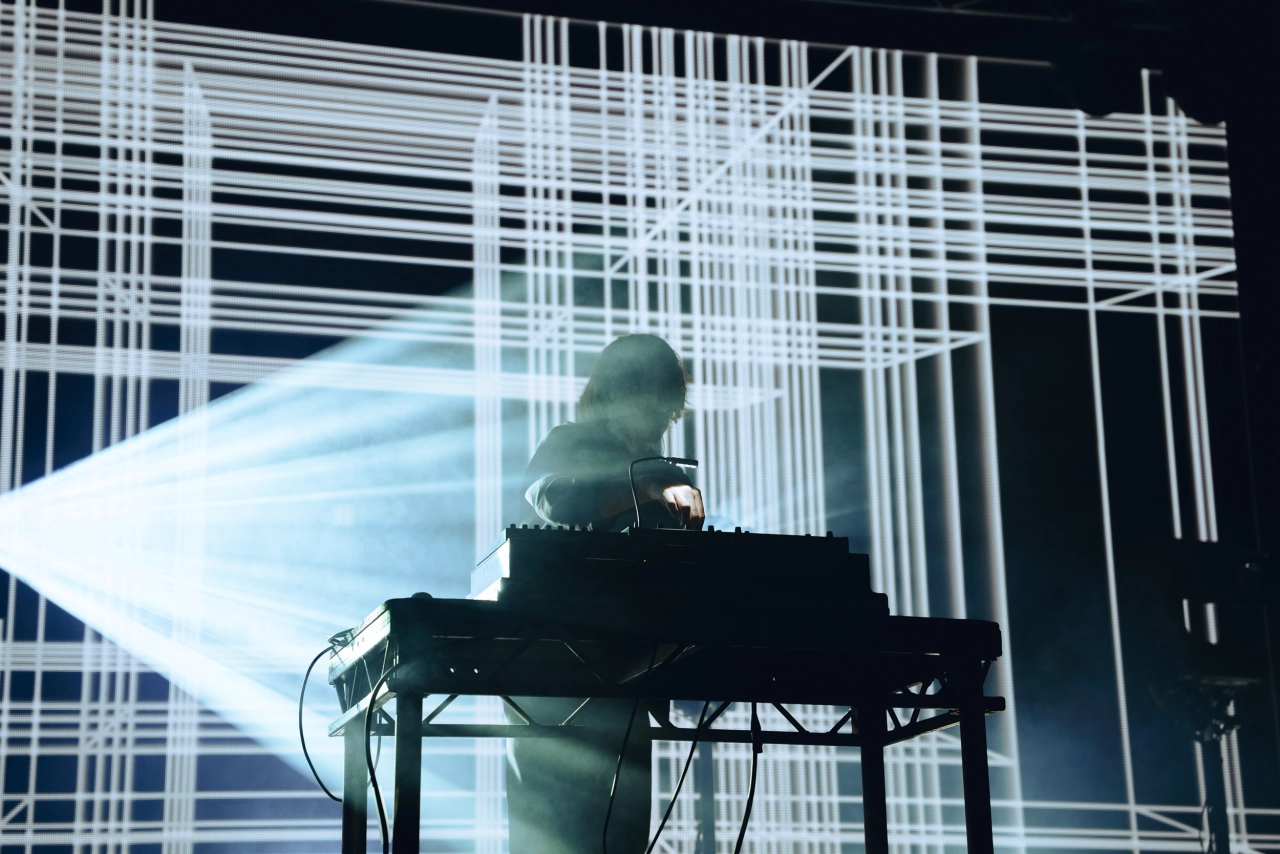 A DJ operates electronic equipment on a raised stand illuminated by a powerful beam of light against a high-contrast geometric backdrop.