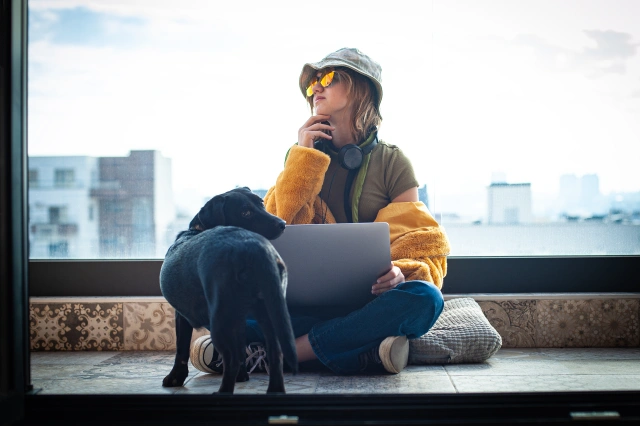 Person sitting by a window with a laptop, wearing a yellow jacket and cap, with a black dog beside them, overlooking a city skyline