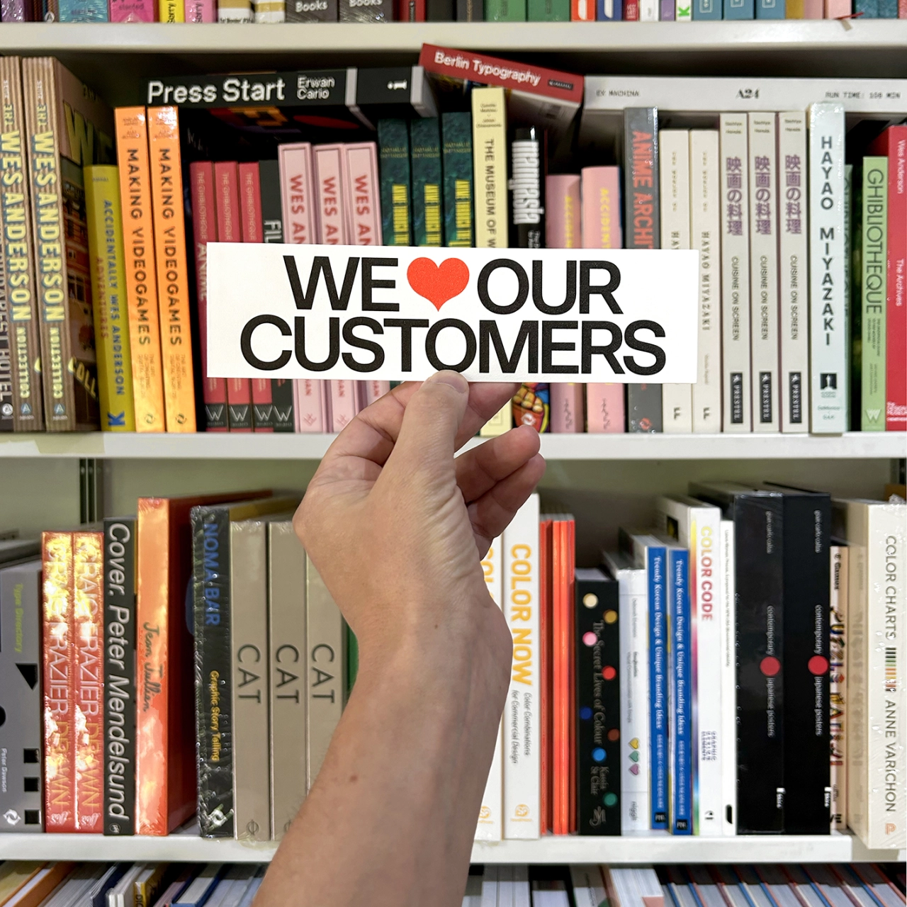 Hand holding a sticker reading "WE LOVE OUR CUSTOMERS" with a red heart, in front of a bookshelf filled with design and typography books