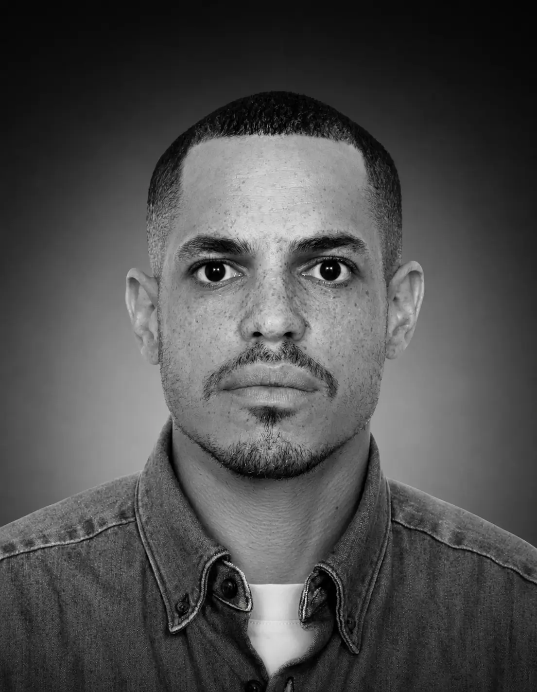 Black and white headshot portrait of a man with short dark hair and facial hair, wearing a denim shirt