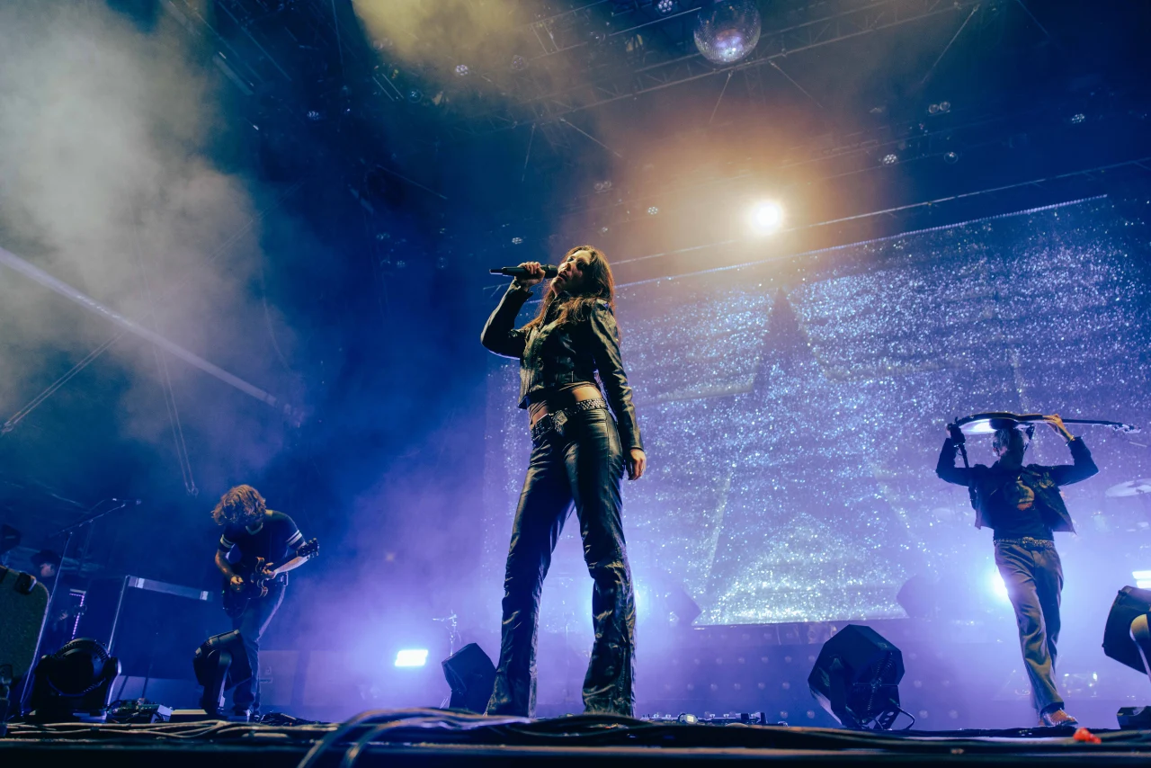 A lead singer stands centered under dramatic stage lighting with her band members playing guitars behind her against a sparkling backdrop.