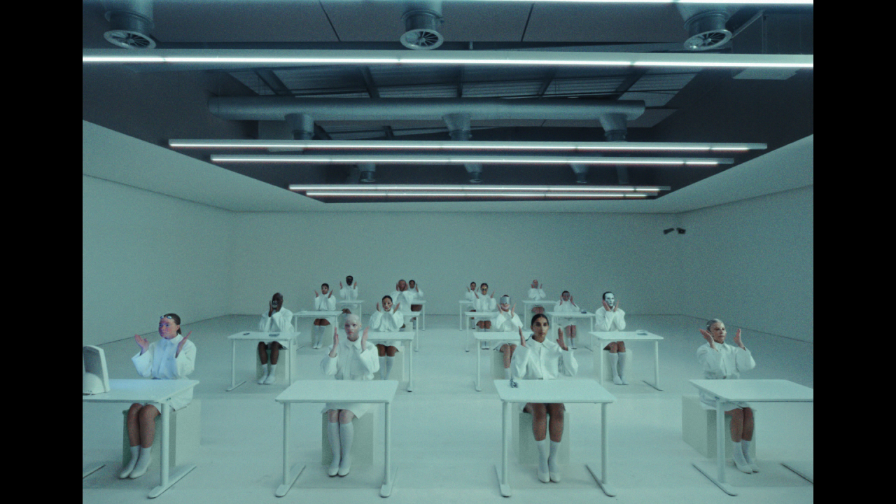 Multiple people in white clothing seated at individual white desks in a minimalist white room with linear ceiling lights