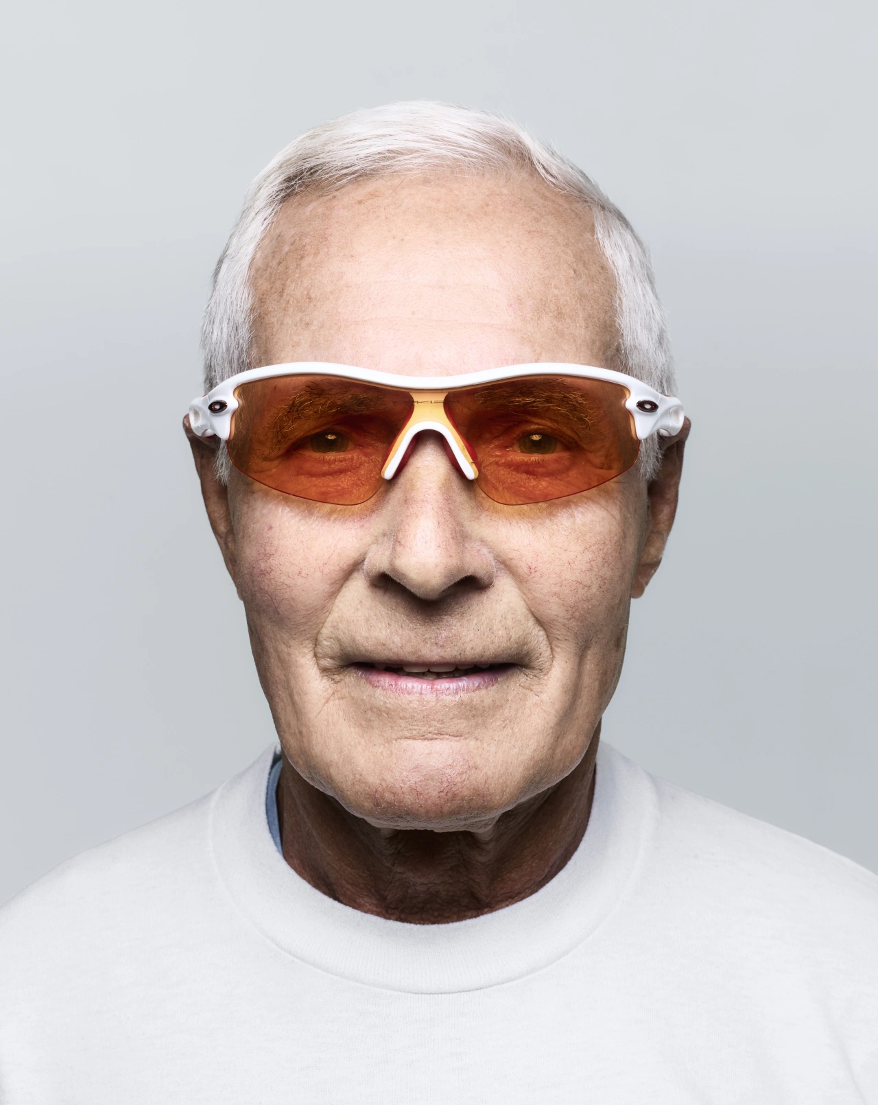 Older man with white hair wearing white-framed sunglasses with amber lenses and a white t-shirt against a grey background