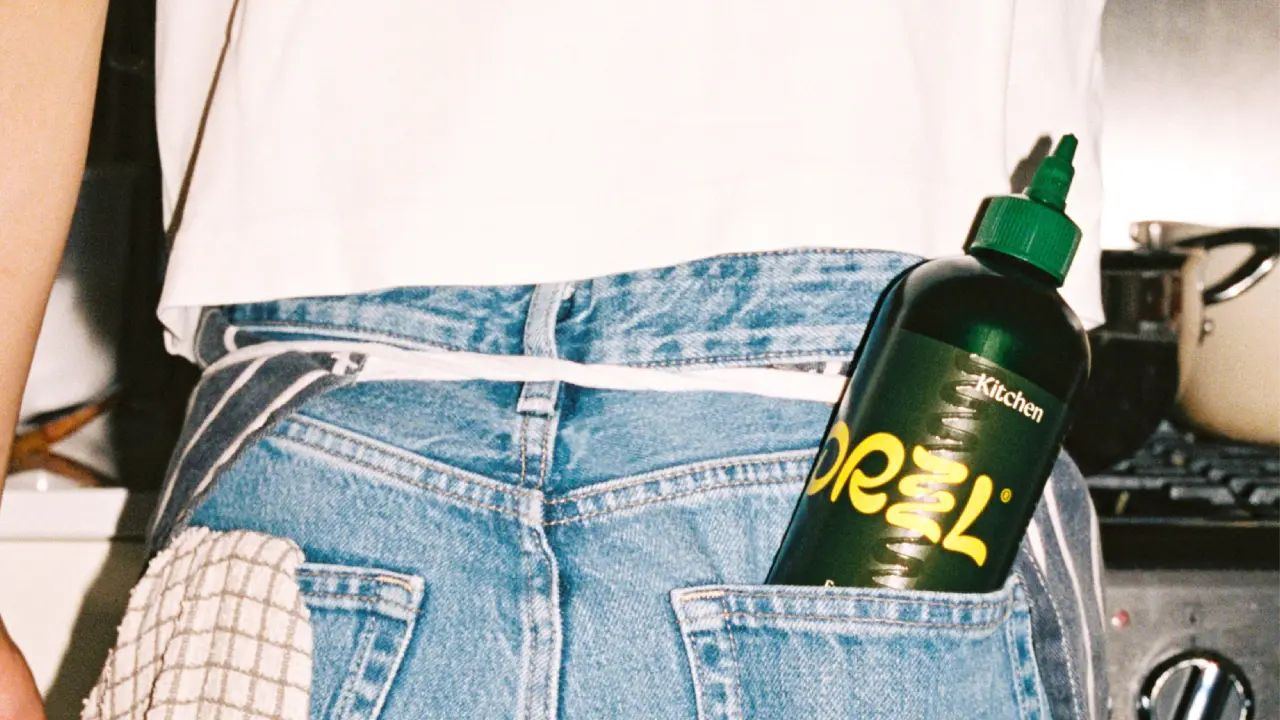 Bottle of Drzzl olive oil with yellow label tucked into the back pocket of blue jeans worn by a person in a white top