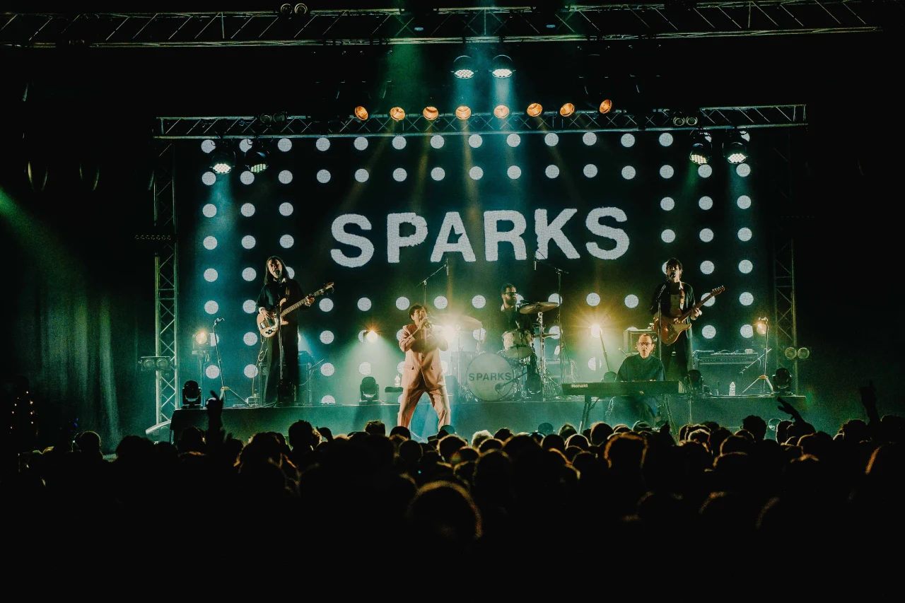 The band Sparks performs on a illuminated stage with a bright grid backdrop displaying their name above a large festival audience.