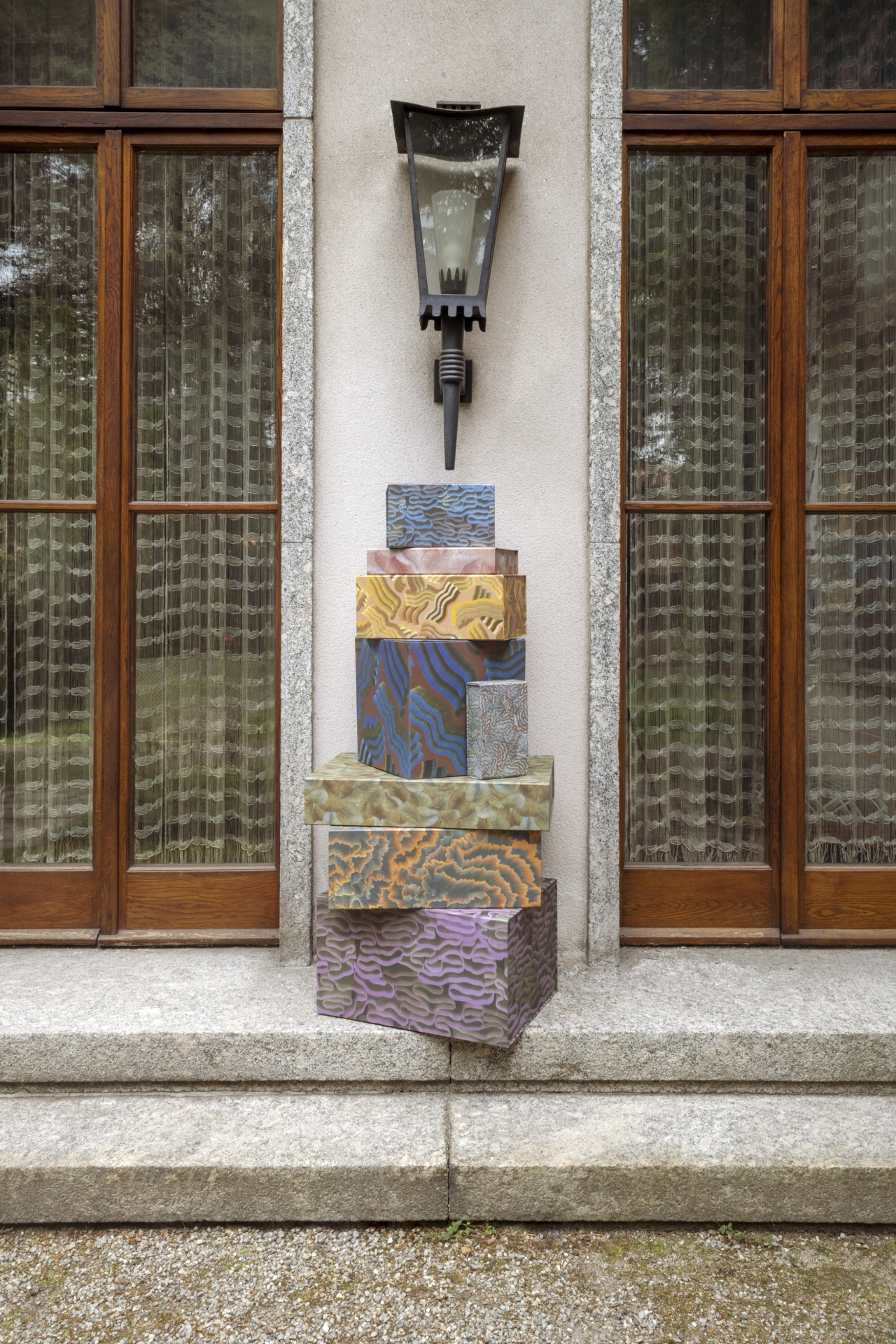 Stacked patterned fabric boxes with a wall-mounted lantern above them, positioned against wooden-framed windows on a white-rendered wall