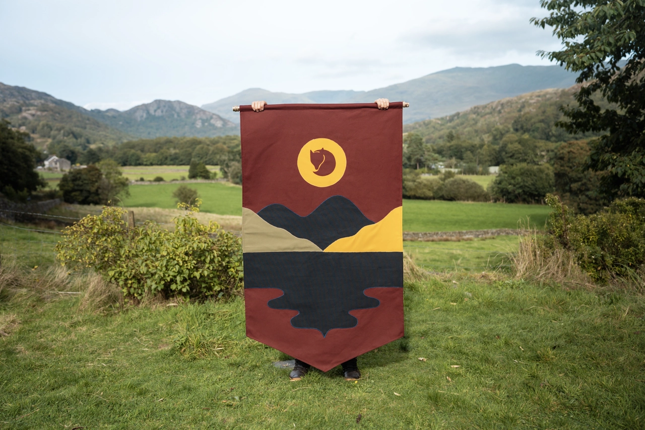 Hand-stitched fabric banner by Miscellaneous Adventures featuring a stylised sun, mountains and forest landscape in red, yellow and black, displayed in a Lake District valley
