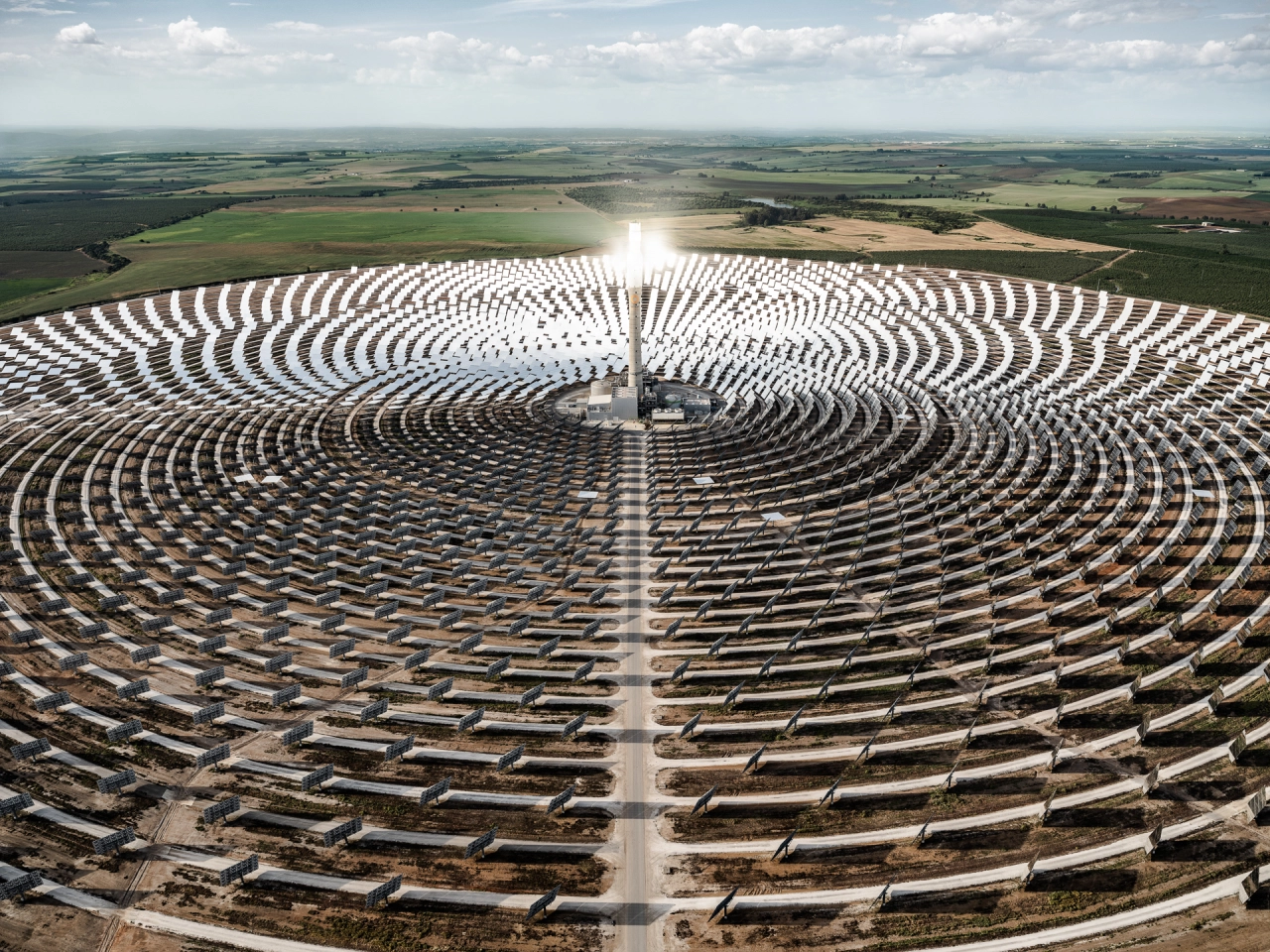 Aerial view of a vast solar thermal power plant with concentric circular rows of mirrors radiating from a central tower beneath cloudy skies, surrounded by agricultural fields