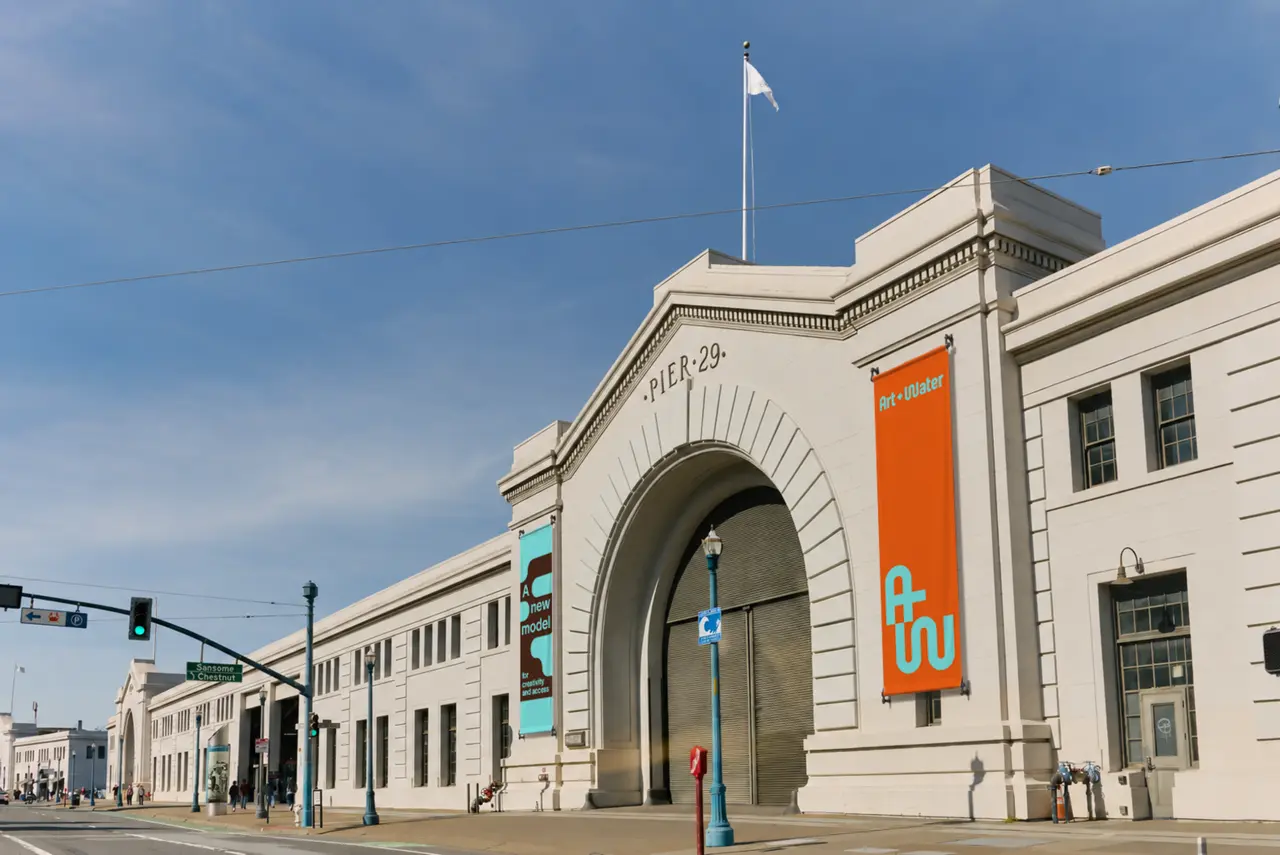 Neoclassical waterfront building with large arched entrance displaying orange and teal banners, under blue sky with flag pole on roofline