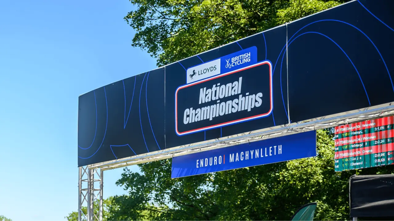 Large outdoor billboard displaying Lloyds British Cycling National Championships branding with blue signage and green foliage behind it