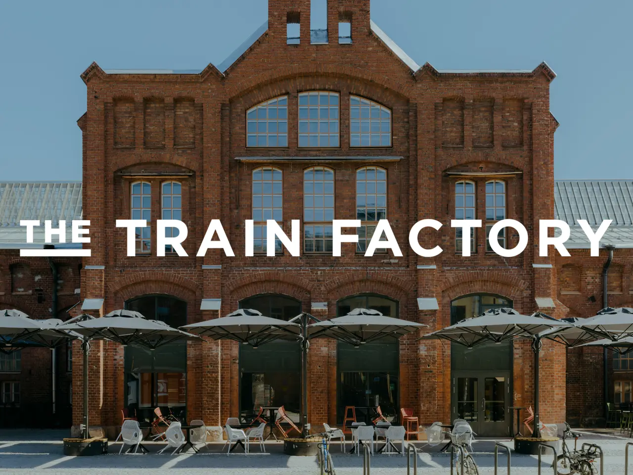 Exterior of The Train Factory, a red brick industrial building in Helsinki with large arched windows and a central gable, featuring a ground-floor restaurant terrace with umbrellas and seating