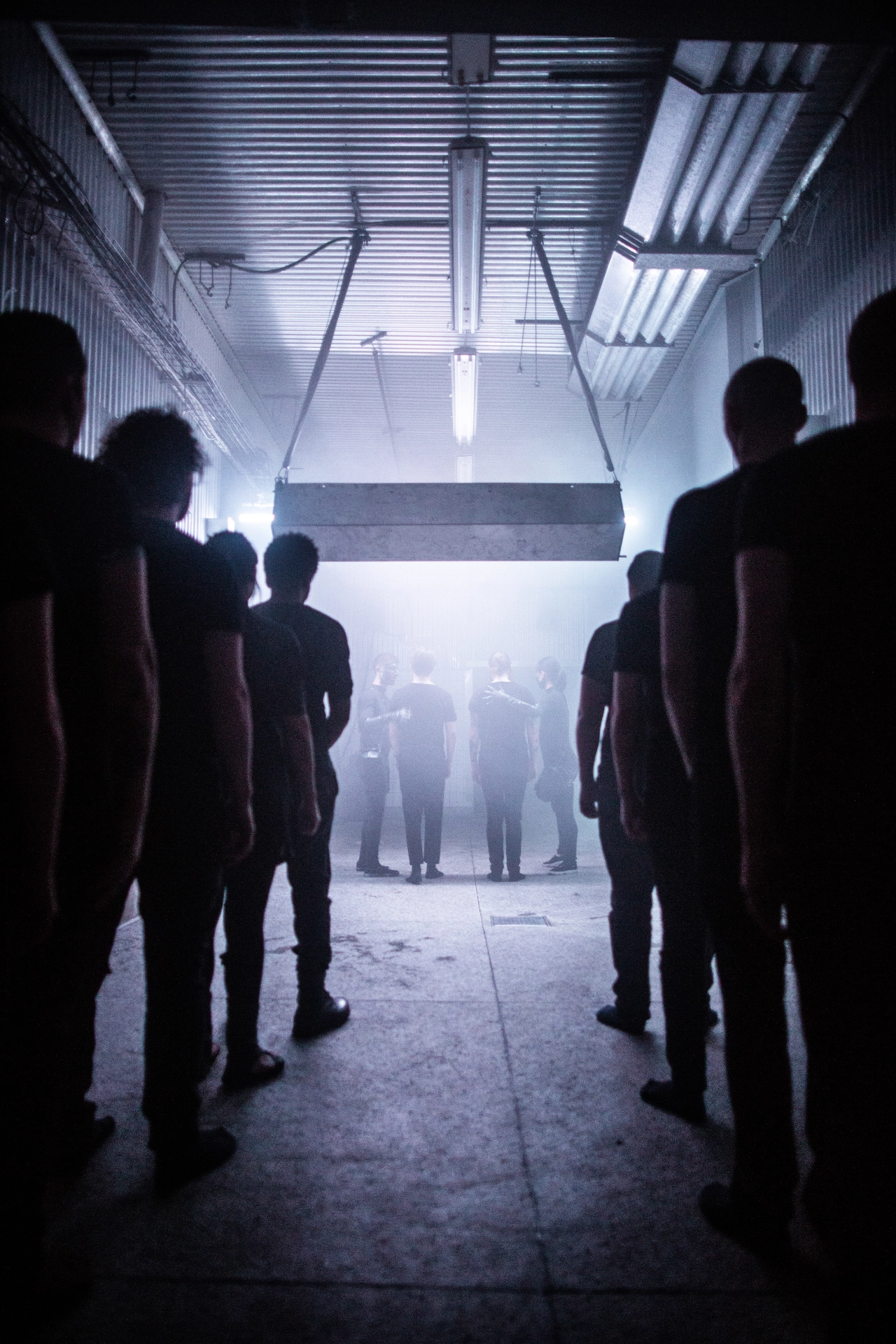 Silhouetted figures stand in a dimly lit industrial warehouse corridor lined with fluorescent lights and corrugated metal walls, facing towards a bright opening ahead