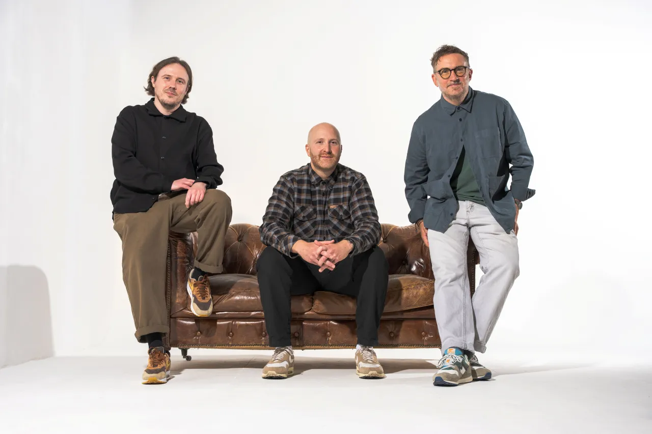 Three men sit on a brown leather sofa against a white background, representing the senior leadership team of commerce practice COMMON following a merger