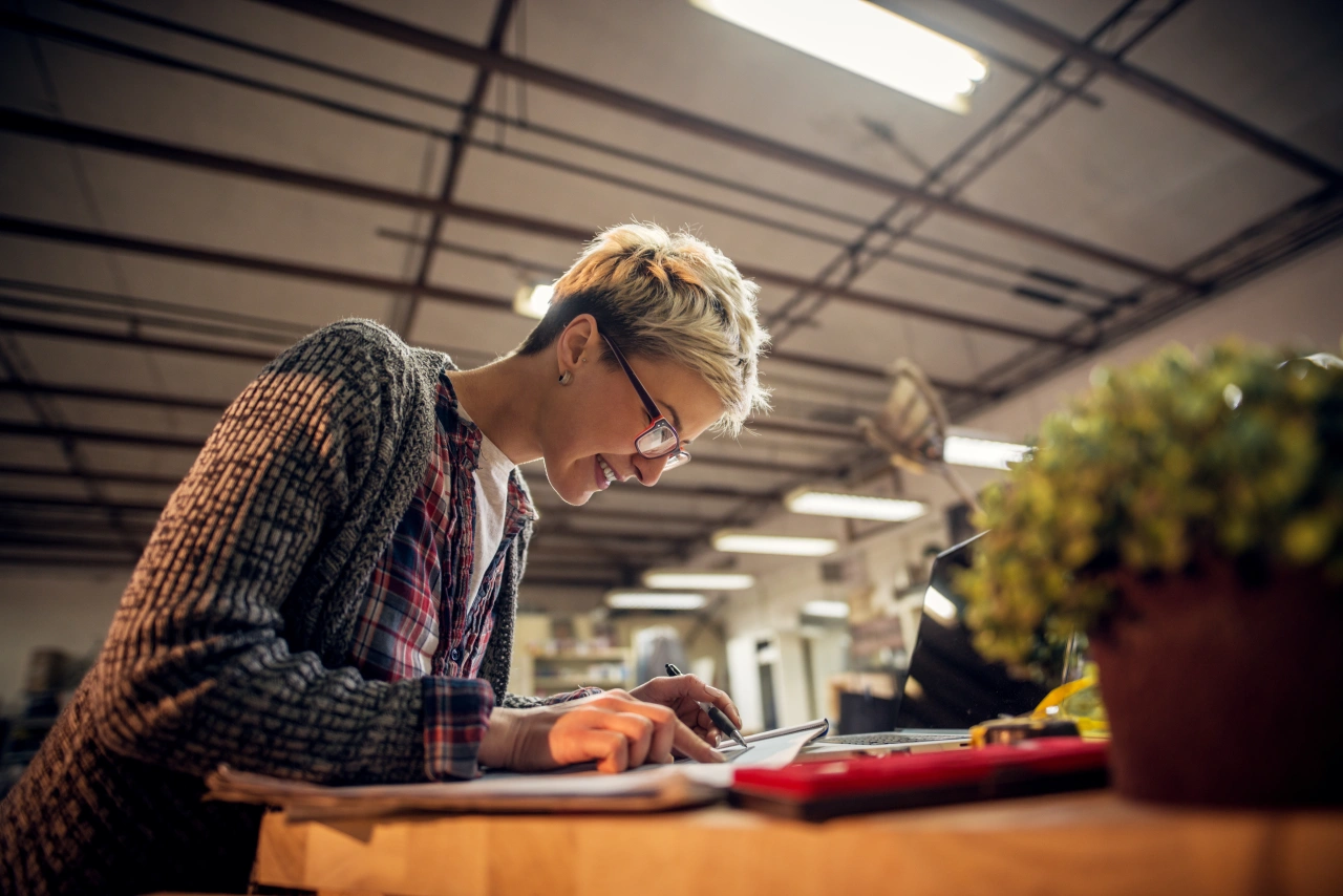 Person with blonde hair wearing glasses and a checked shirt works at a wooden desk in a bright industrial workspace with potted plants nearby