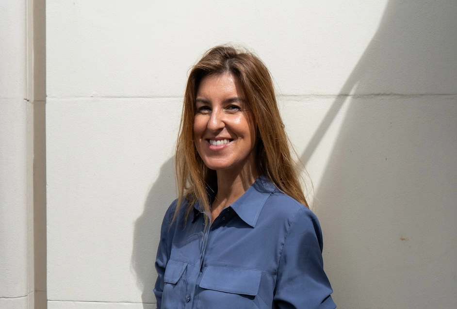 Woman with long brown hair wearing a blue shirt, smiling at the camera against a white wall with shadow detail