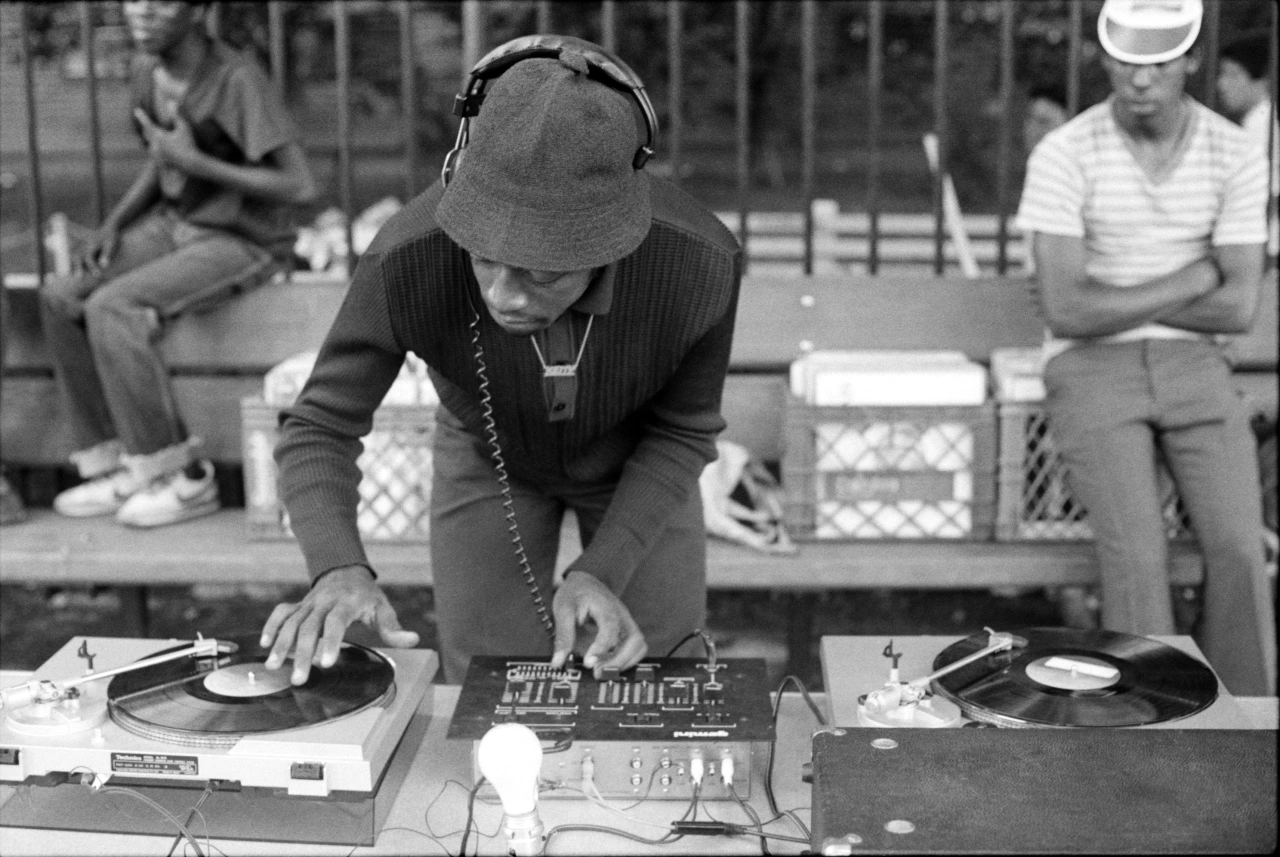 DJ K Slay performs at turntables wearing headphones and a baseball cap in a black and white photograph by Henry Chalfant documenting 1980s New York street culture