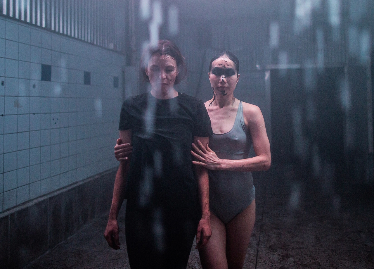 Two women in dark swimwear stand together in a tiled room as part of the Chapter Five/KAIHOGYO immersive art experience