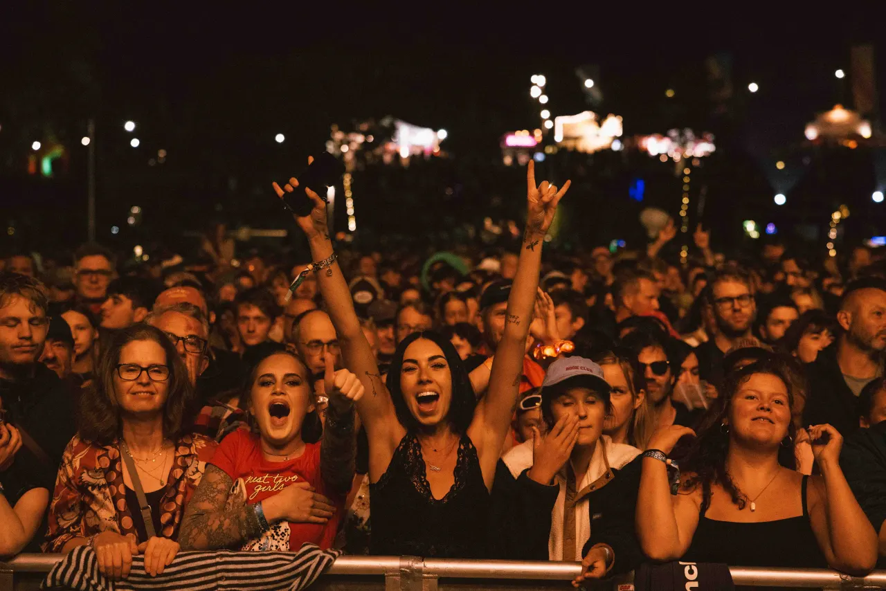 Festival audience members cheer and raise their hands at the front barrier during an evening outdoor concert.