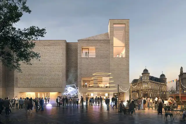 Modern brick building with illuminated windows and tall tower element, with people gathered in front plaza at dusk, historic buildings visible in background