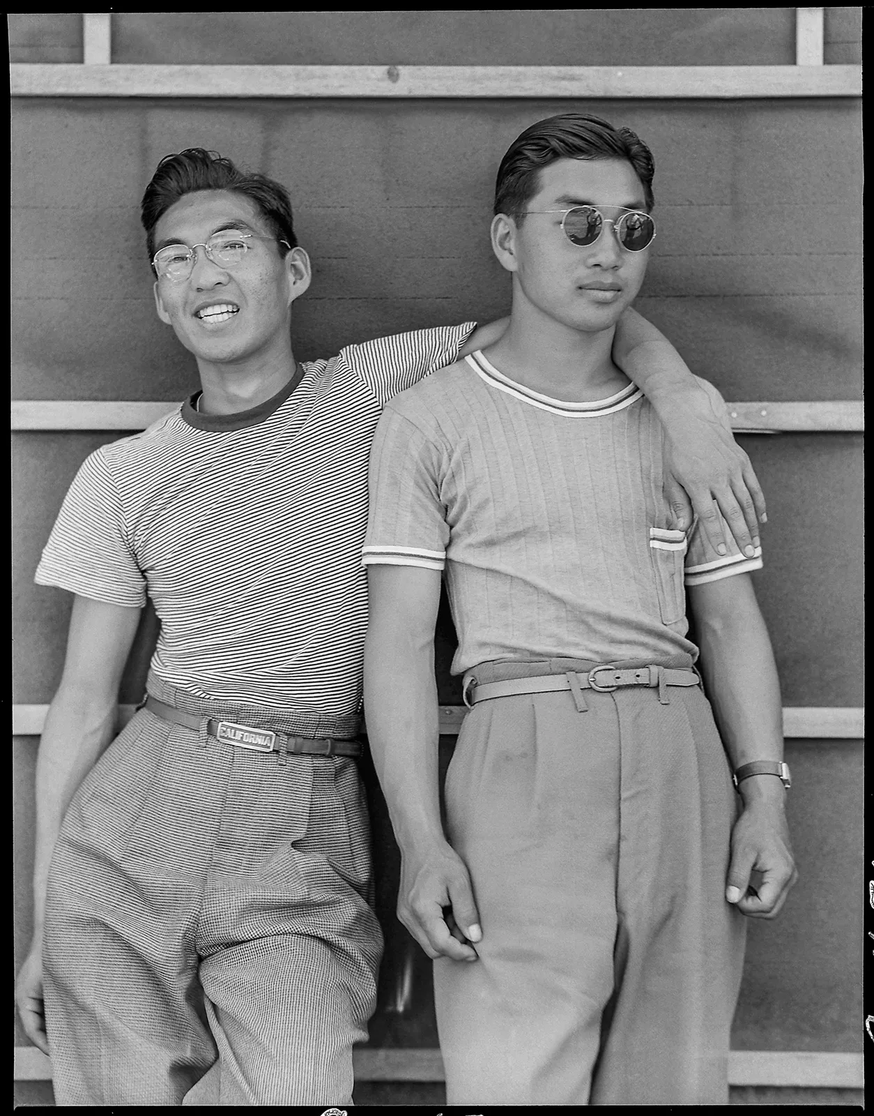 Two young men in 1940s casual clothing stand against a wall at Manzanar internment camp, one wearing glasses and striped shirt, the other in round sunglasses and plain shirt