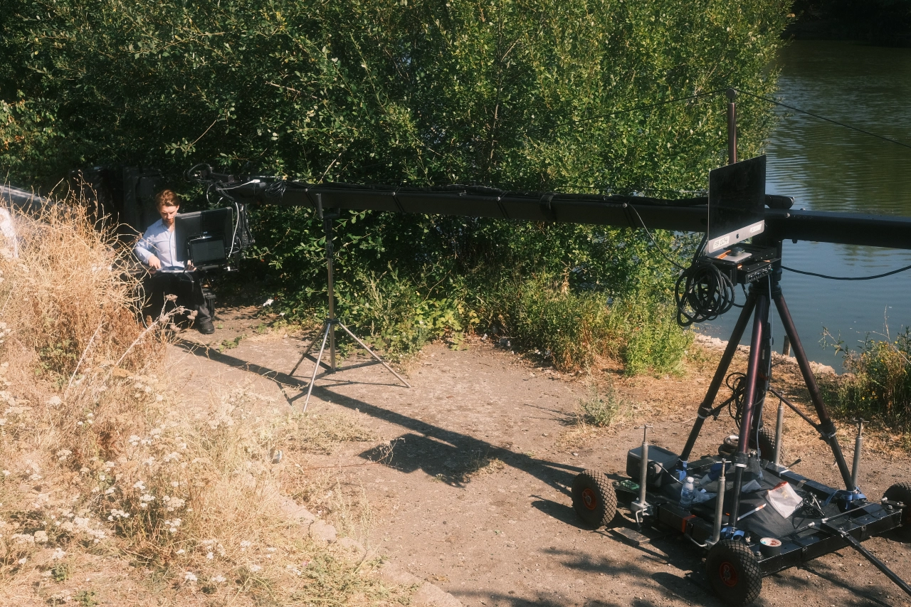 A makeshift film crane with a long black arm extends over a sandy riverbank, with a camera mounted on one end and an iMac computer visible on the other end