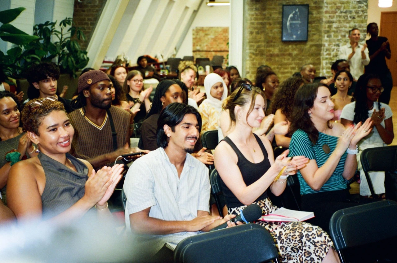 A diverse audience seated in rows applauds at an indoor event with exposed brick walls and pendant lighting