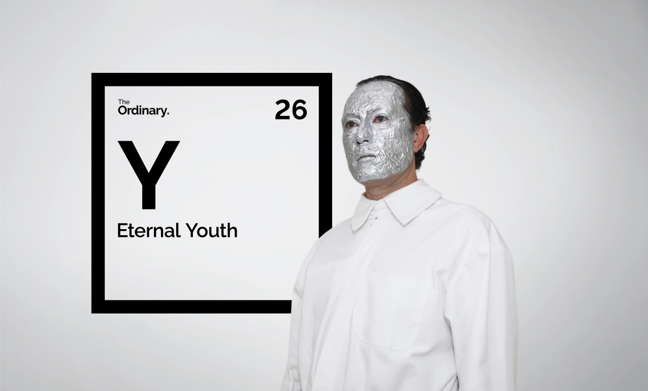 Man wearing white shirt with face covered in white clay mask standing next to periodic table element card reading "Y Eternal Youth
