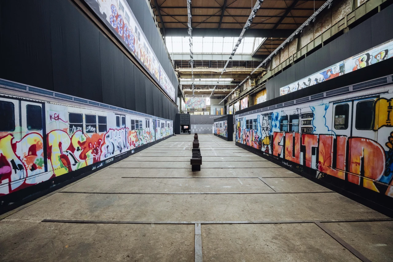 Two colourfully graffitied trains parked facing each other inside an industrial warehouse space with skylights overhead