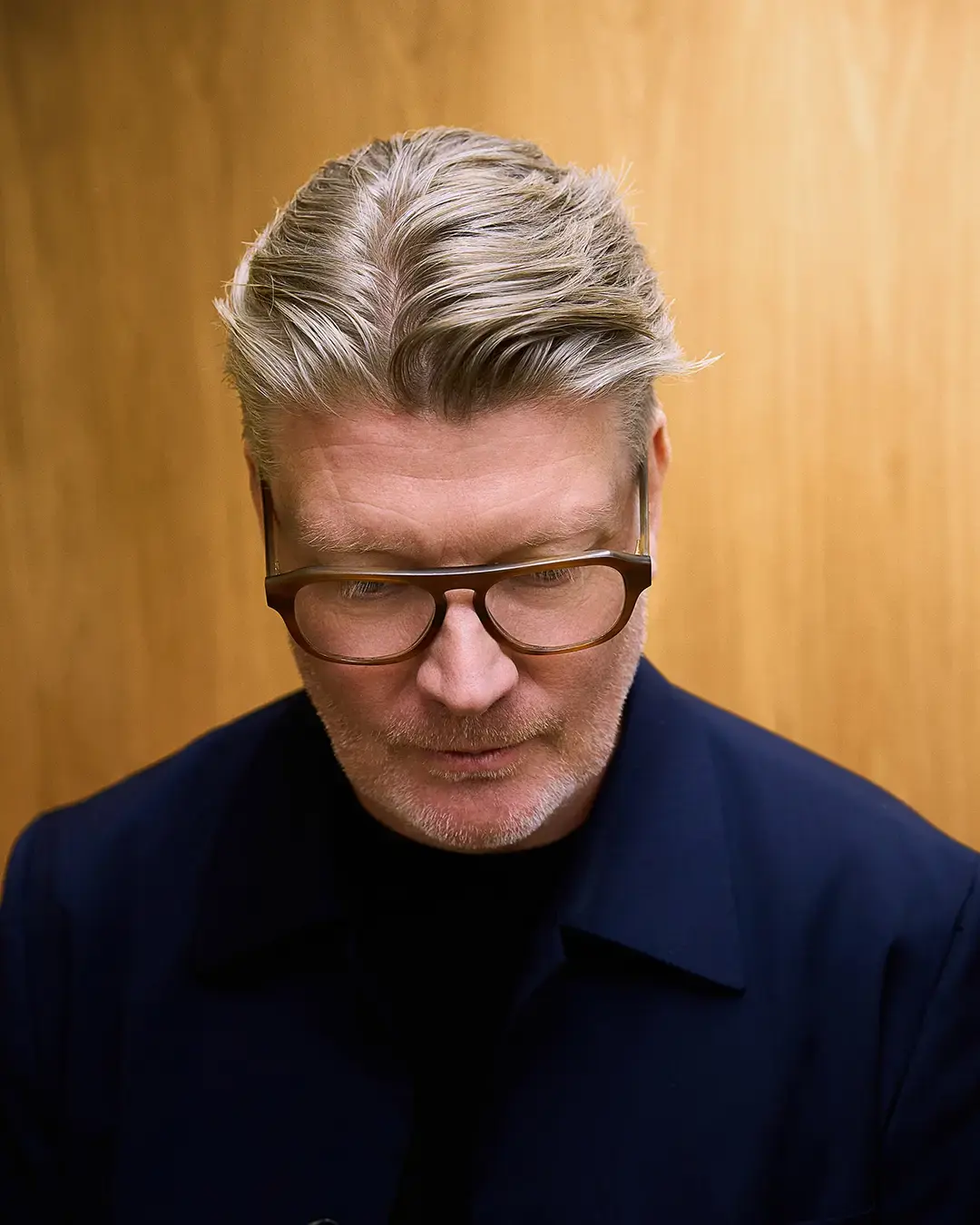 Simon Dixon, co-founder of DixonBaxi, wearing glasses and a dark navy shirt against a mustard-coloured background