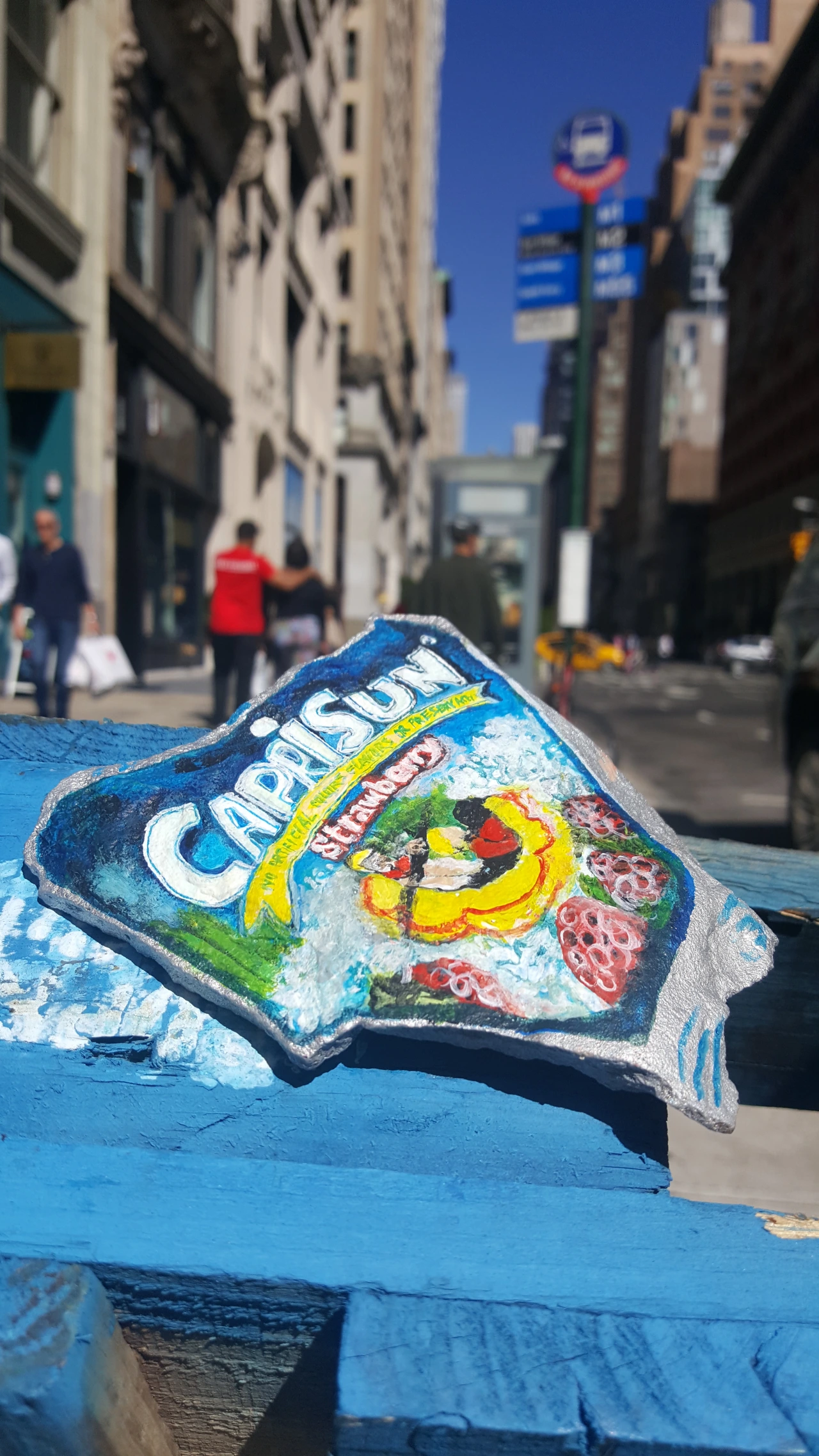 Painted rock featuring a Capri Sun juice pouch design with yellow stripes and illustrated strawberries, displayed on a blue surface with a city street in the background