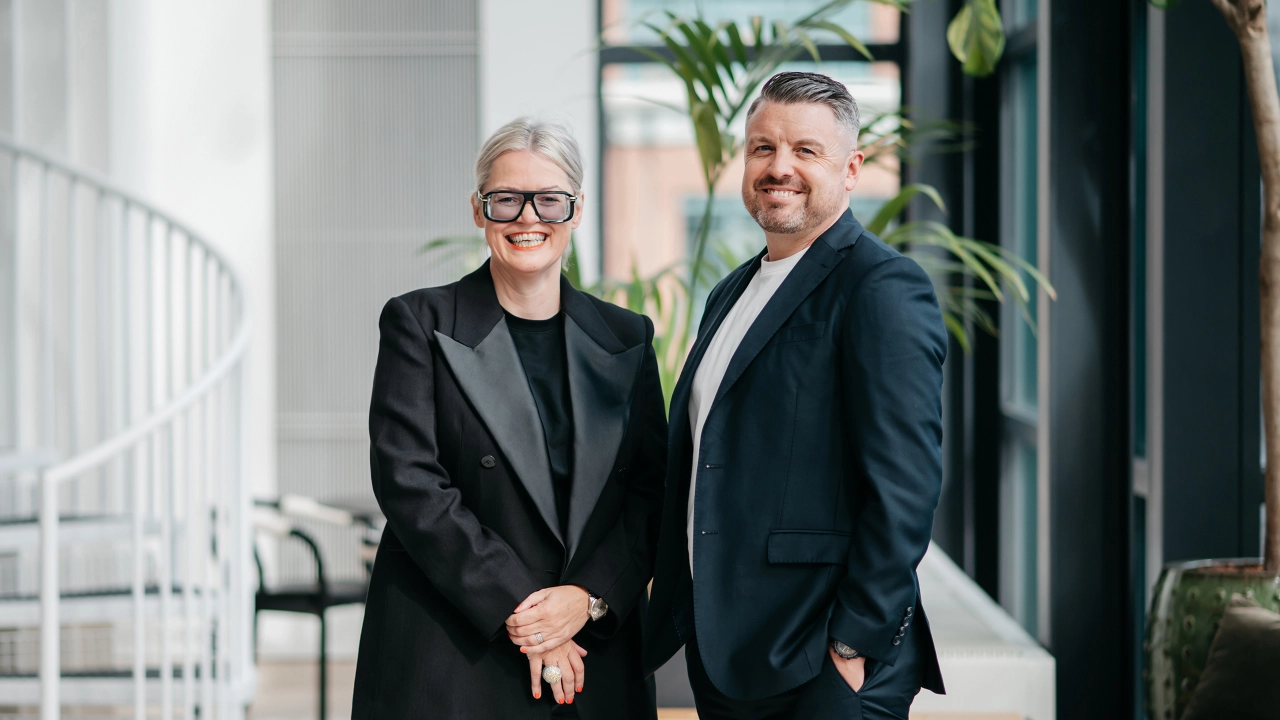 Cerys James and Chris Falconer smile at the camera whilst shaking hands in a modern office corridor with plants and white railings visible behind them