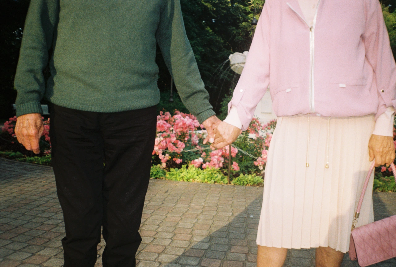 Two people holding hands while walking past pink flowers in a garden, wearing green and pink cardigans