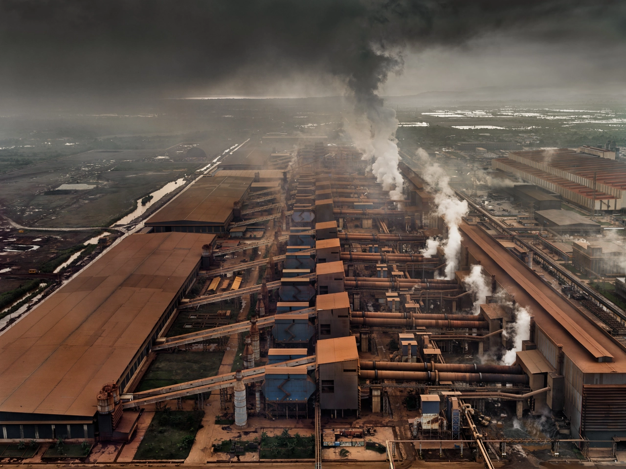 Aerial view of an industrial mining complex with multiple rectangular extraction ponds in orange and blue, surrounded by farmland, with white smoke rising from processing facilities