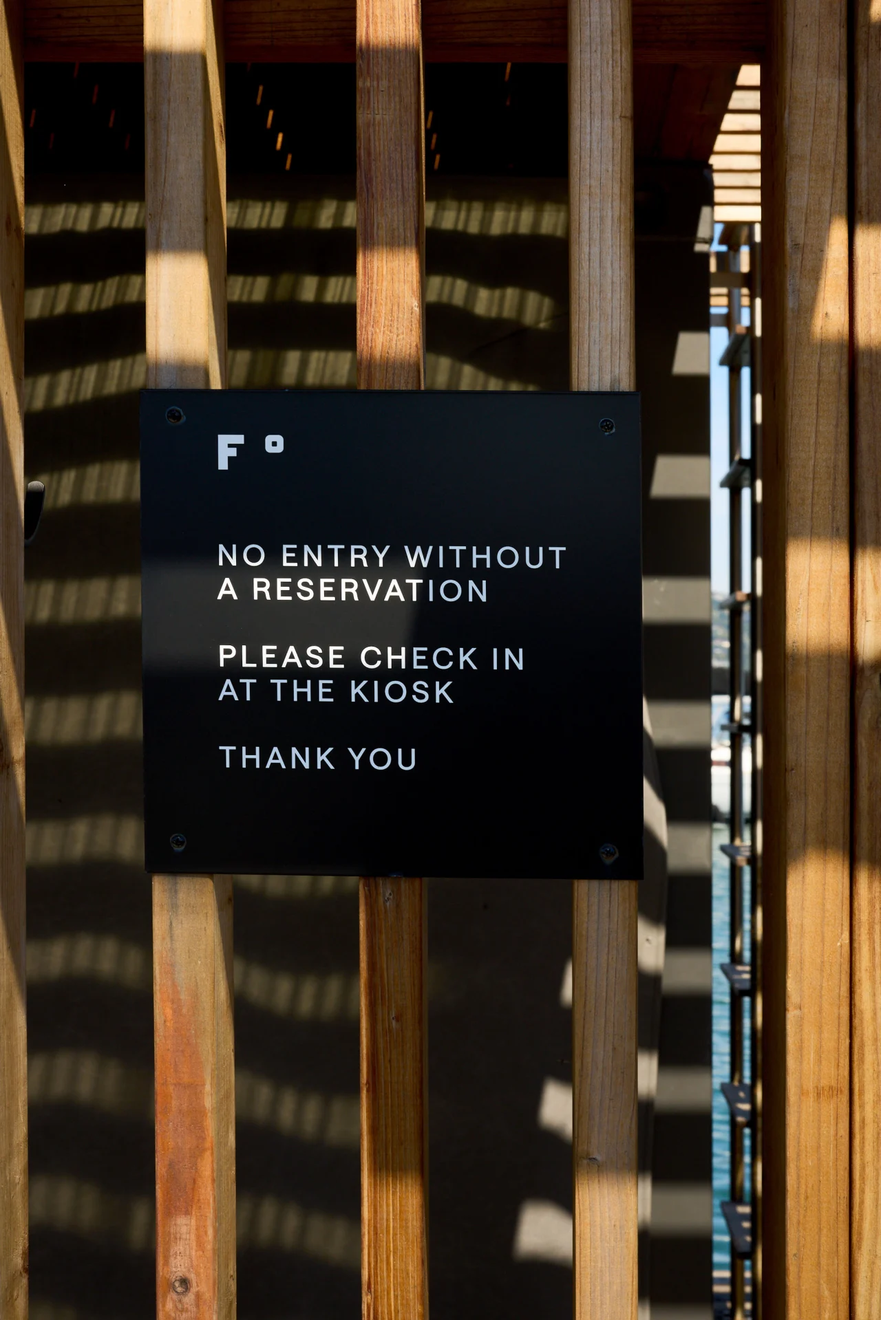 Black sign mounted on wooden gate reading "F°" logo, "NO ENTRY WITHOUT A RESERVATION", "PLEASE CHECK IN AT THE KIOSK" and "THANK YOU", with striped shadows cast across wooden slats behind it