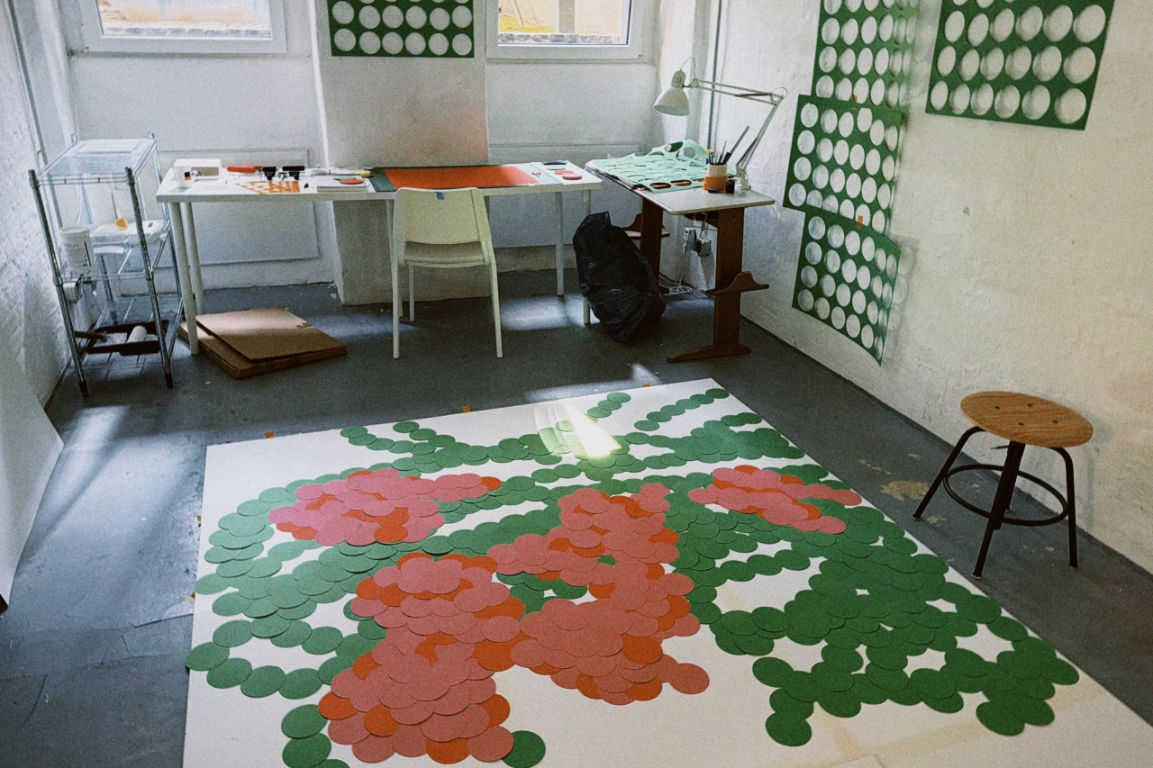 Artist Ayong Son's studio workspace with hand-cut paper flowers in coral and green covering the floor, desk with materials, and egg displays on walls