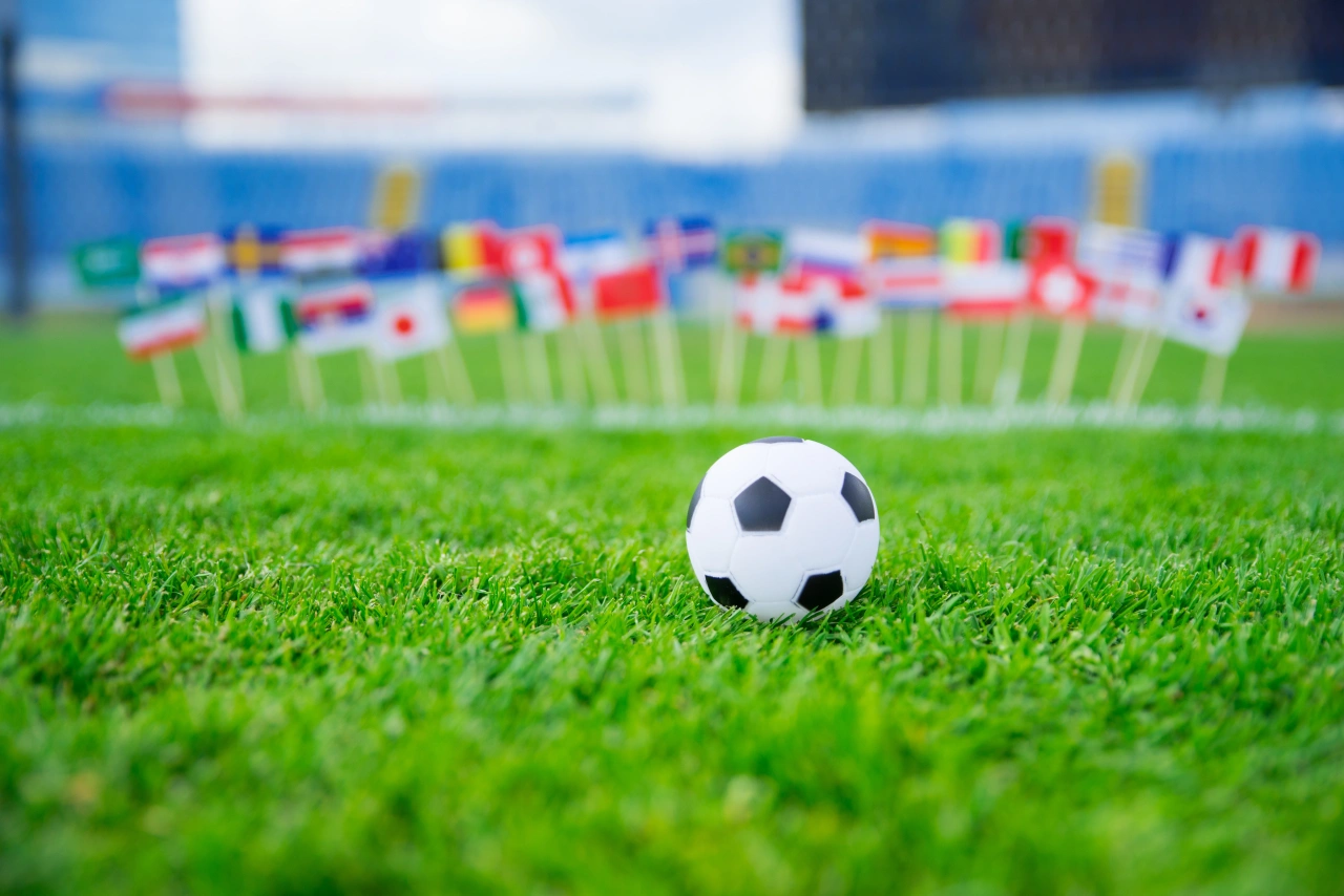Football on grass pitch with international flags blurred in background stadium seating