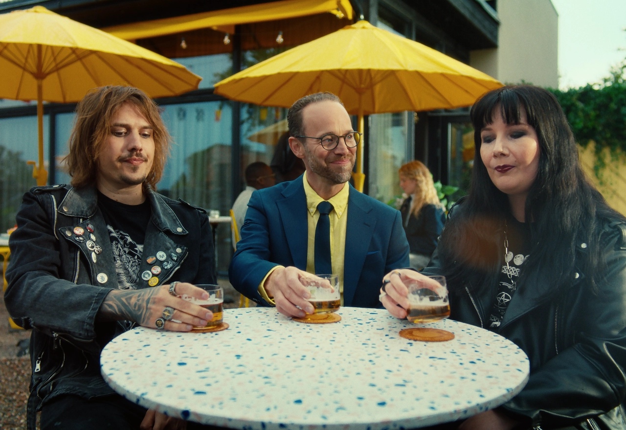 Three people sit at a speckled outdoor table beneath yellow umbrellas, holding sandwiches and drinks at the Introvert Travel Agency pop-up in London