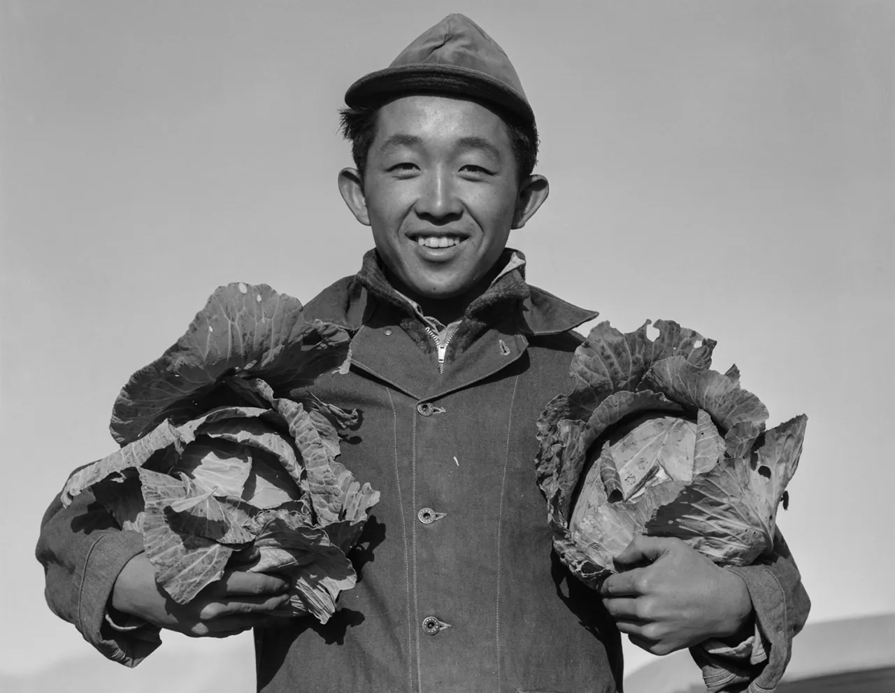 A young man wearing a cap and jacket smiles at the camera while holding two large heads of cabbage, one in each hand