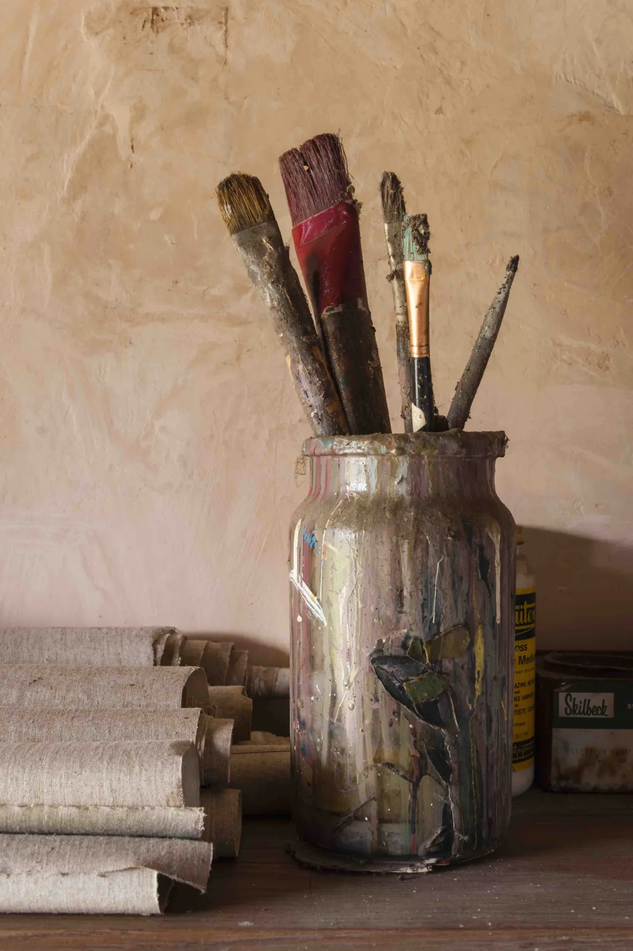 Worn paintbrushes in various sizes and colours stand upright in a paint-splattered glass jar beside rolled paper and art supplies in Sidney Nolan's studio