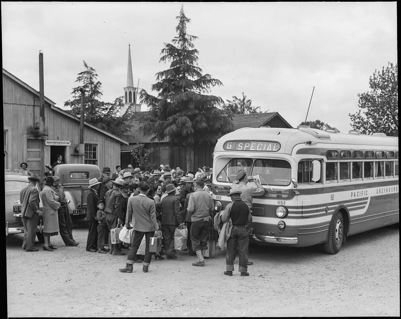 A crowd of Japanese American evacuees and officials gather around a bus marked "Special" at Manzanar internment camp, with a church steeple visible behind them