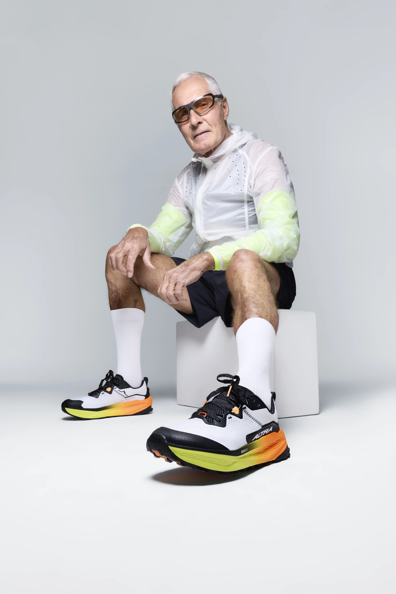 Older man wearing glasses and high-visibility running jacket crouches on a white block, displaying colourful running trainers with neon yellow and orange accents