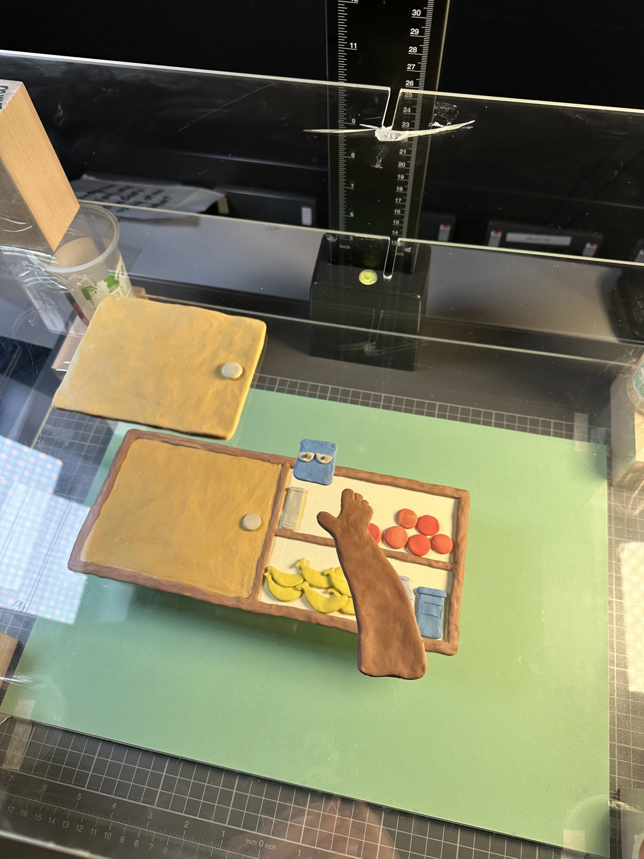 Overhead view of a clay animation multiplane set with wooden countertops, yellow clay items, and miniature fruit arranged on a green surface beneath adjustable glass plates and measurement guides
