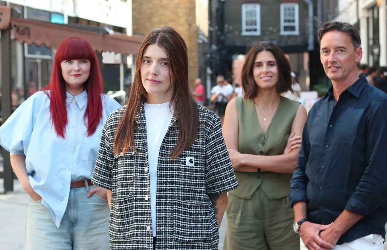 Four people standing together outdoors in front of urban buildings, including Natalie Prout in a checkered shirt in the centre