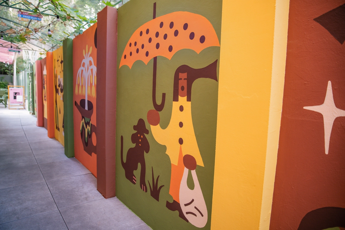 Colourful mural panels lining a corridor feature a figure with an umbrella sheltering a small silhouette in the rain, painted in warm earth tones and geometric shapes