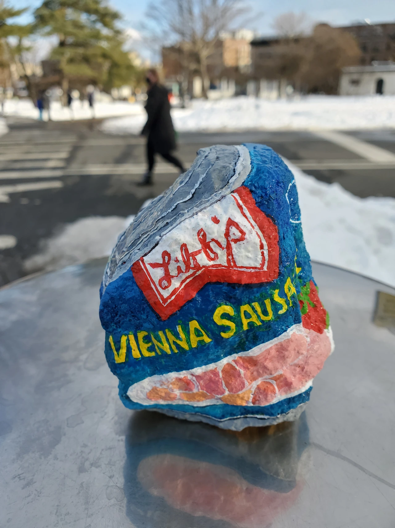 A painted rock depicting a Libby's Vienna Sausage tin sits on a snowy surface with a blurred street scene and buildings behind it